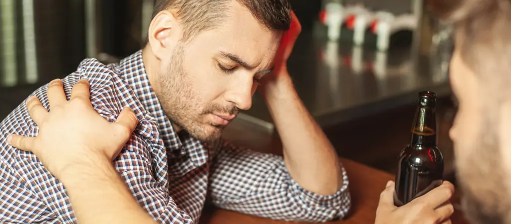 Agg&oacute;d&oacute; f&eacute;rfi &uuml;l az asztaln&aacute;l alkoholos itallal, mik&ouml;zben egy m&aacute;sik szem&eacute;ly t&aacute;mogat&oacute;n meg&eacute;rinti a v&aacute;ll&aacute;t, szeml&eacute;ltetve a szerencsej&aacute;t&eacute;k f&uuml;ggős&eacute;g megelőz&eacute;se sor&aacute;n fontos emberi seg&iacute;ts&eacute;get